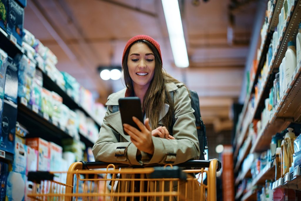 Happy woman using mobile app on her phone while shopping in supermarket.